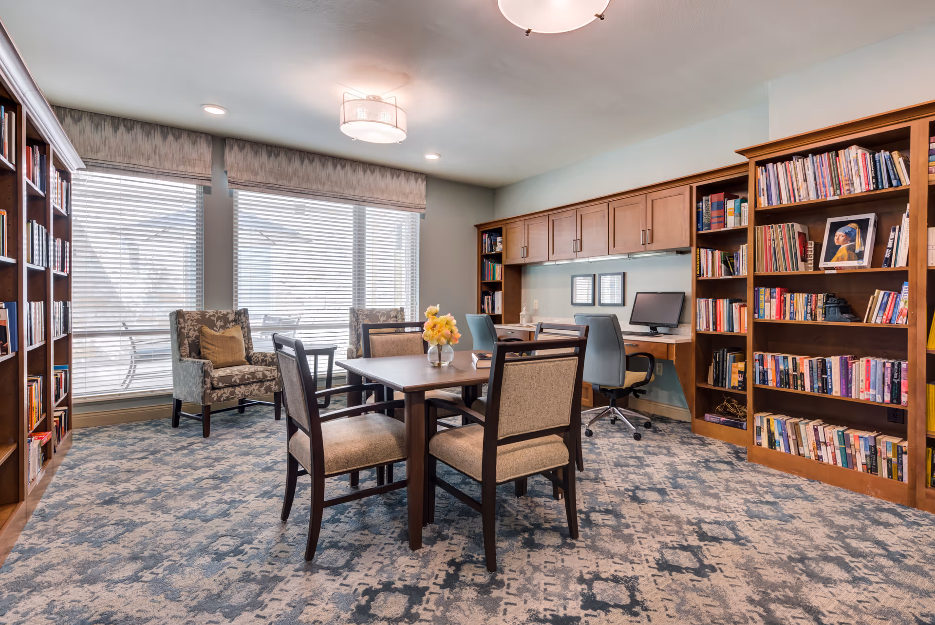 A well-lit library or study room with large windows covered by blinds. The room features wooden bookshelves filled with books, a central table with four chairs, two upholstered armchairs near the window, and a built-in desk area with two office chairs and a computer. The carpet has a patterned design and the ceiling has modern light fixtures.