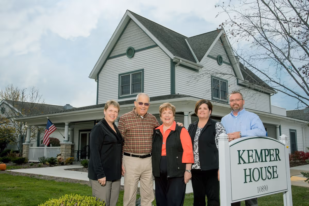 Five adults standing outside a white two-story building with green trim, smiling at the camera. They are positioned near a sign that reads 'Kemper House 10890'. The building has a porch with columns and an American flag is visible on the left side.
