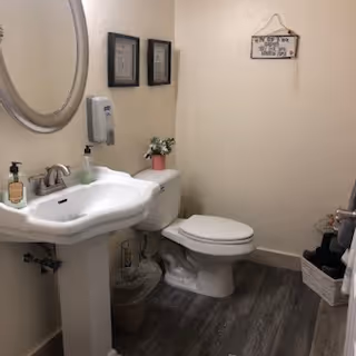 A small bathroom with a white pedestal sink, a round mirror above it, a soap dispenser mounted on the wall, and a bottle of hand soap on the sink. Next to the sink is a white toilet with a small plant on the tank. The floor has wood-like vinyl flooring, and there are framed pictures on the wall above the toilet. A wastebasket is placed between the sink and toilet.