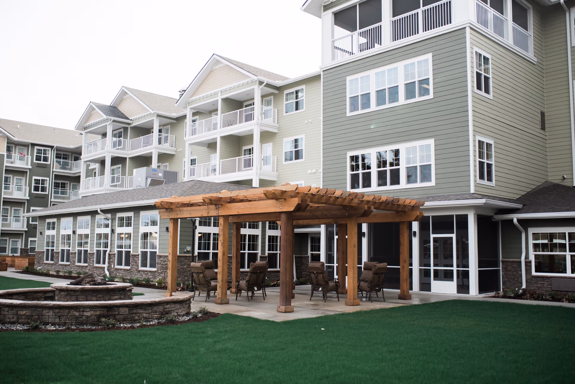 Outdoor seating area with a wooden pergola and cushioned chairs on a concrete patio, adjacent to a multi-story residential building with balconies and numerous windows, surrounded by green grass and a stone fire pit.