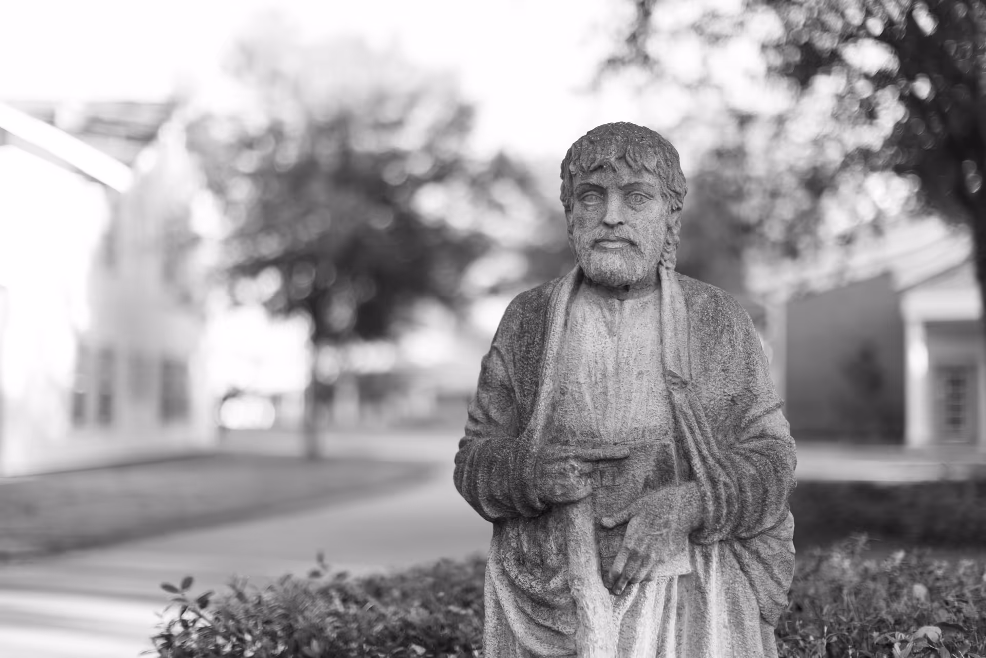 Black and white photo of a stone statue of a bearded man wearing a robe, standing outdoors with blurred trees and buildings in the background.