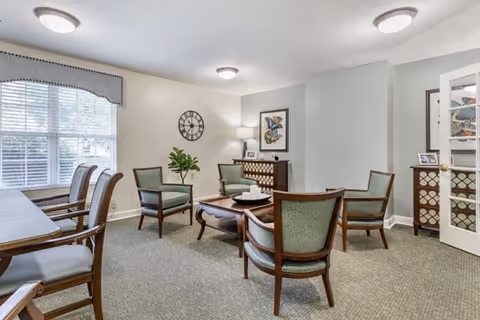 A cozy sitting area with four wooden armchairs upholstered in green fabric arranged around a wooden coffee table. There is a sideboard with a lamp and framed artwork on the wall above it. A large window with blinds and a valance lets in natural light. The room has neutral-colored walls and carpeted flooring.