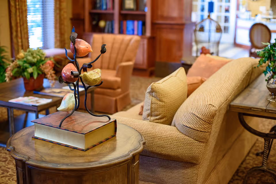 A cozy living room area with a beige textured sofa adorned with cushions, a wooden side table holding a decorative sculpture of colorful birds on branches, an armchair in the background, a bookshelf filled with books, and a coffee table with a flower arrangement and magazines.
