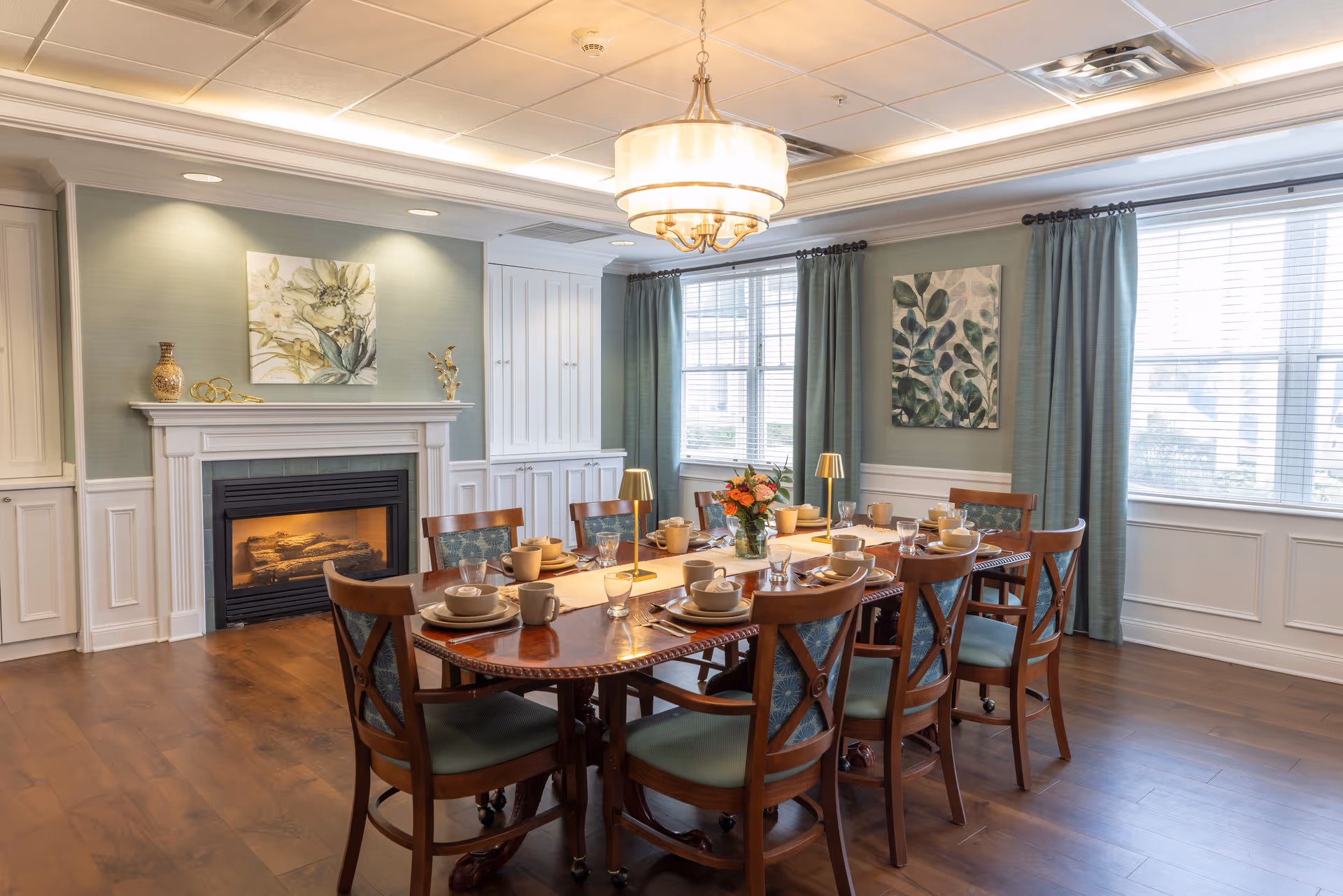 A bright dining room with a wooden table set for a meal, chairs, a fireplace, chandelier, and large windows with green curtains.