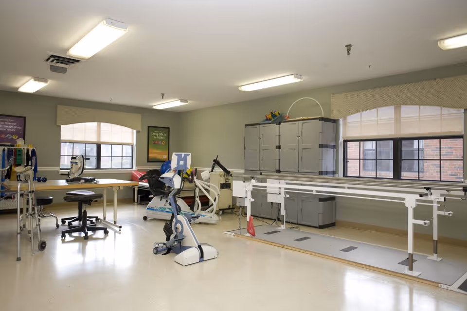 A spacious physical therapy room with exercise equipment including a stationary bike, parallel bars for walking practice, a walker, and therapy tables. The room has large windows with beige valances, light green walls, and bright overhead lighting.