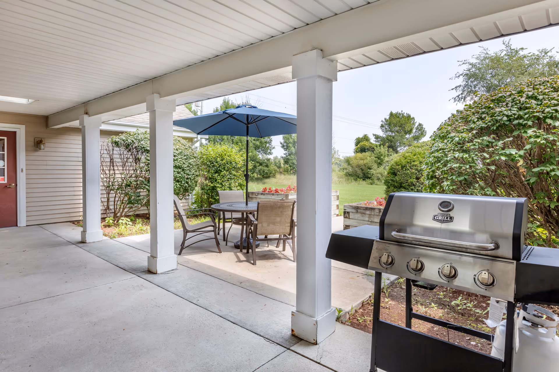 Covered outdoor patio area with white pillars, a round table with four chairs, a blue umbrella, and a stainless steel grill. There are bushes and greenery surrounding the patio and an open grassy area beyond.