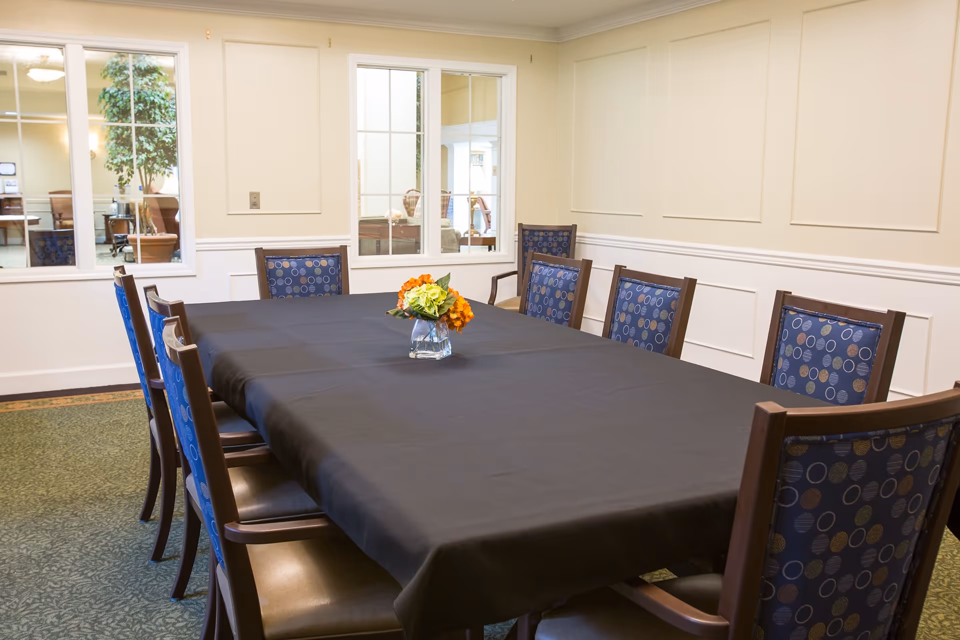 A dining room with a long rectangular table covered with a black tablecloth, surrounded by wooden chairs with blue patterned cushions. A small glass vase with orange and green flowers is placed in the center of the table. The room has cream-colored walls with white wainscoting and windows looking into an adjacent room with a large potted plant.