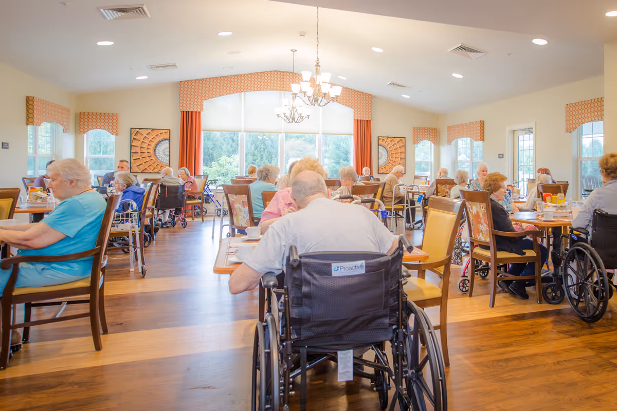 A bright dining room in a senior living facility with elderly residents seated at tables, some in wheelchairs, enjoying a meal. The room has large windows with orange curtains, wooden floors, and chandeliers hanging from the ceiling.