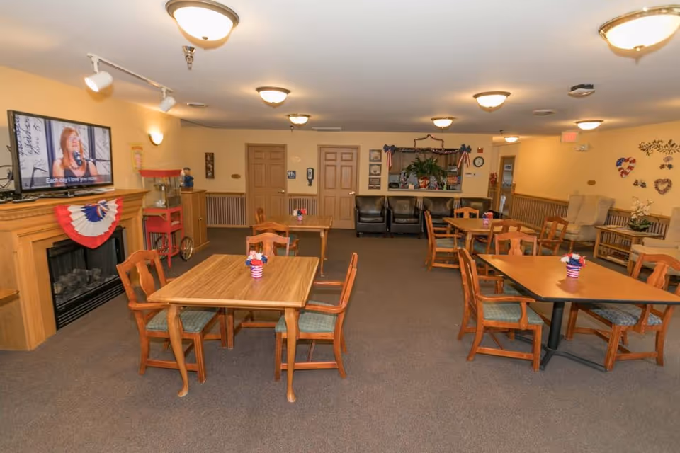 A communal dining room with several wooden tables and chairs arranged neatly. Each table has a small patriotic-themed centerpiece. The room features a fireplace with a patriotic bunting decoration, a television mounted above it displaying a woman singing, and a popcorn machine nearby. The walls are decorated with various patriotic and floral decorations. There are also two black armchairs and a counter with plants and additional decorations in the background.