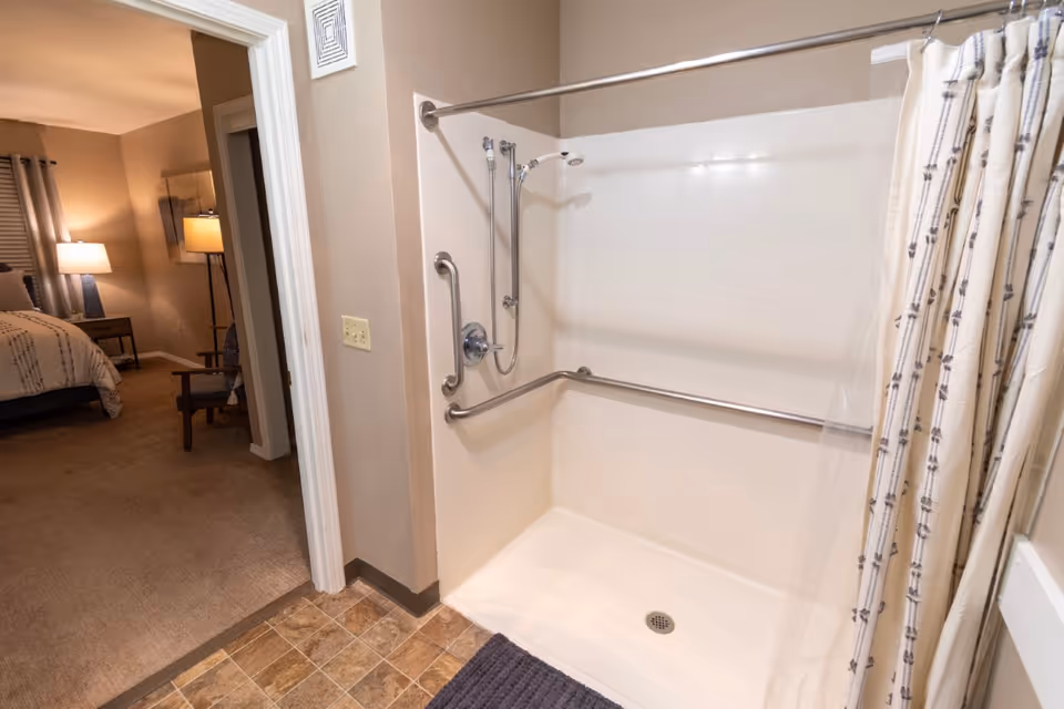 A bathroom shower area with grab bars and a handheld showerhead, adjacent to a bedroom with a bed, nightstand, lamp, and chair visible through an open doorway.
