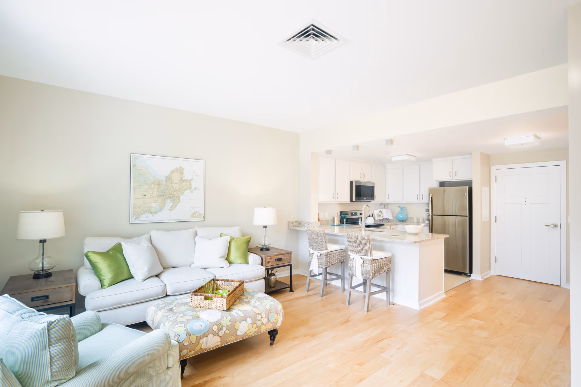 Bright and airy living room and kitchen area with light wood flooring. The living room features a white sofa with green and white pillows, a patterned ottoman, a striped armchair, and two side tables with lamps. The kitchen has white cabinets, a granite countertop island with two wicker bar stools, stainless steel appliances including a refrigerator and microwave, and a white door in the background.