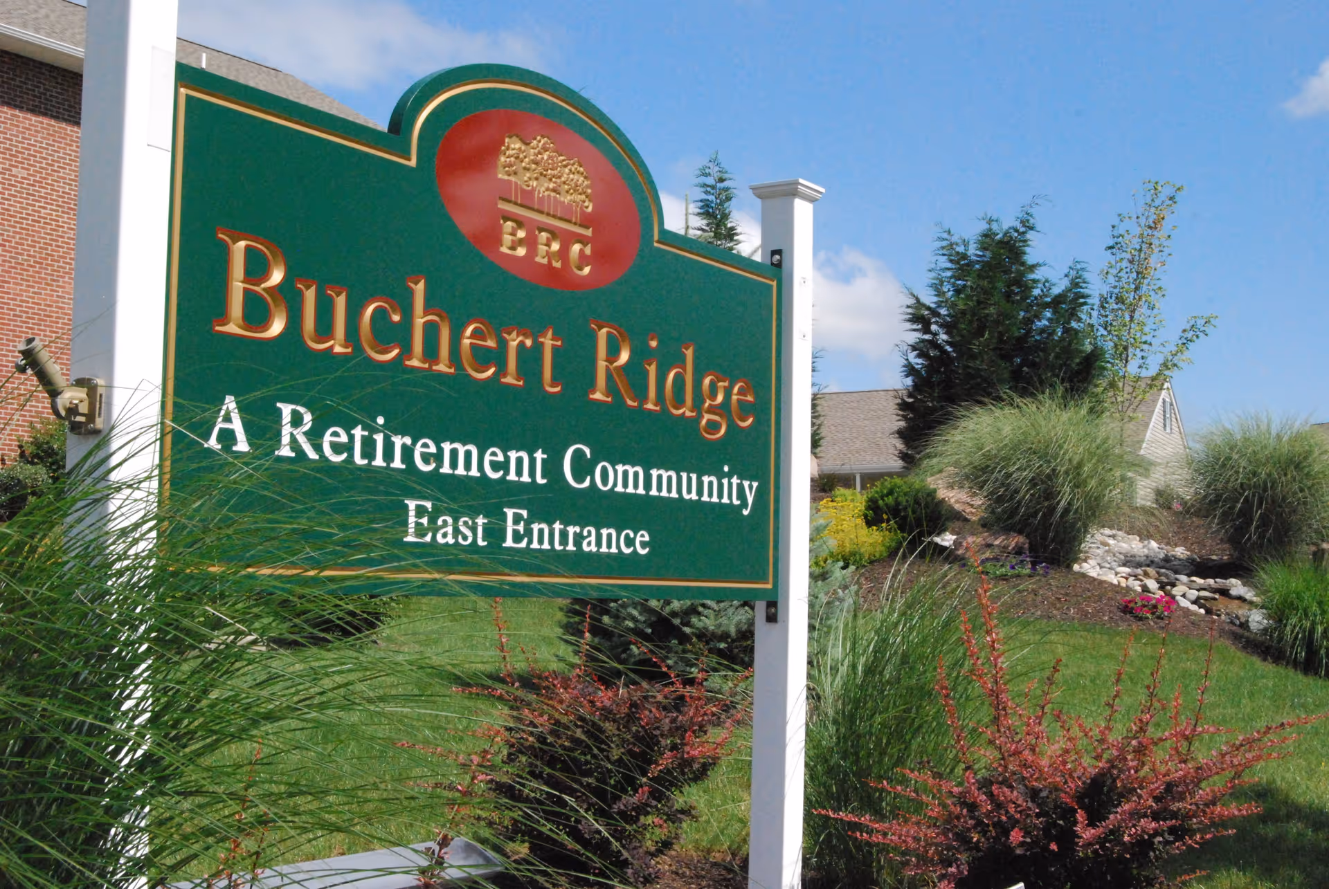 Green and gold sign for Buchert Ridge, a retirement community, marking the East Entrance with landscaping and a building in the background under a blue sky.