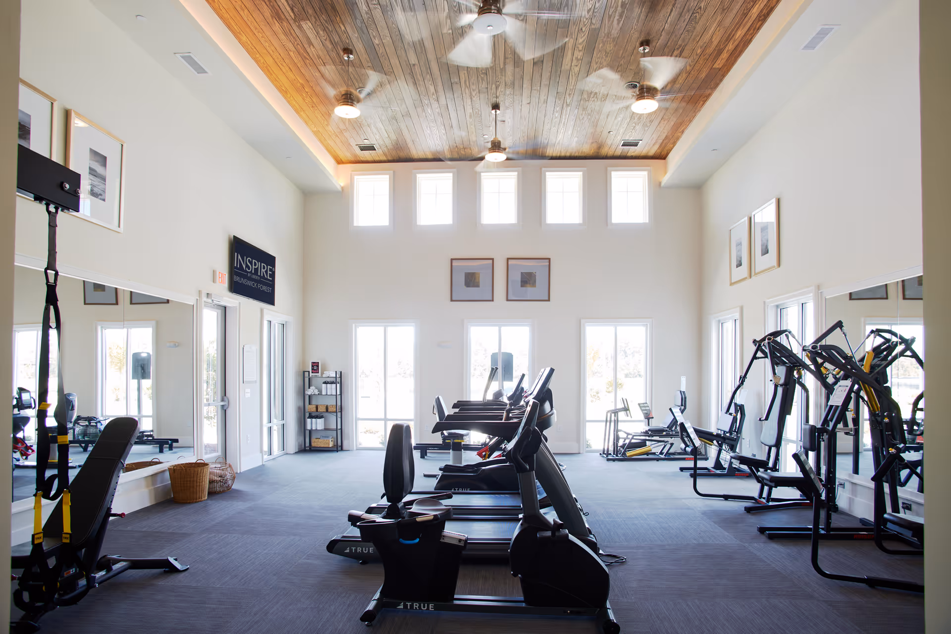 Bright and spacious fitness room with exercise equipment including treadmills, stationary bikes, and weight machines. The room has large windows letting in natural light, a wooden ceiling with ceiling fans, and mirrors on the walls. A sign on the wall reads 'INSPIRE Brunswick Forest'.