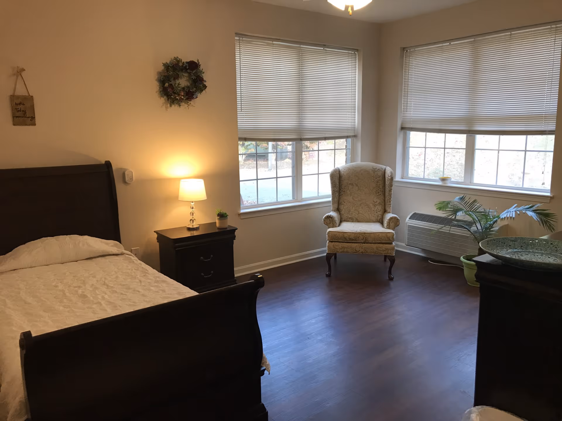A senior living bedroom with a dark wooden bed frame and white bedding, a nightstand with a lamp and small plant, a decorative wreath on the wall, a beige upholstered armchair near two large windows with blinds, a potted plant, and a heating/cooling unit beneath the windows.