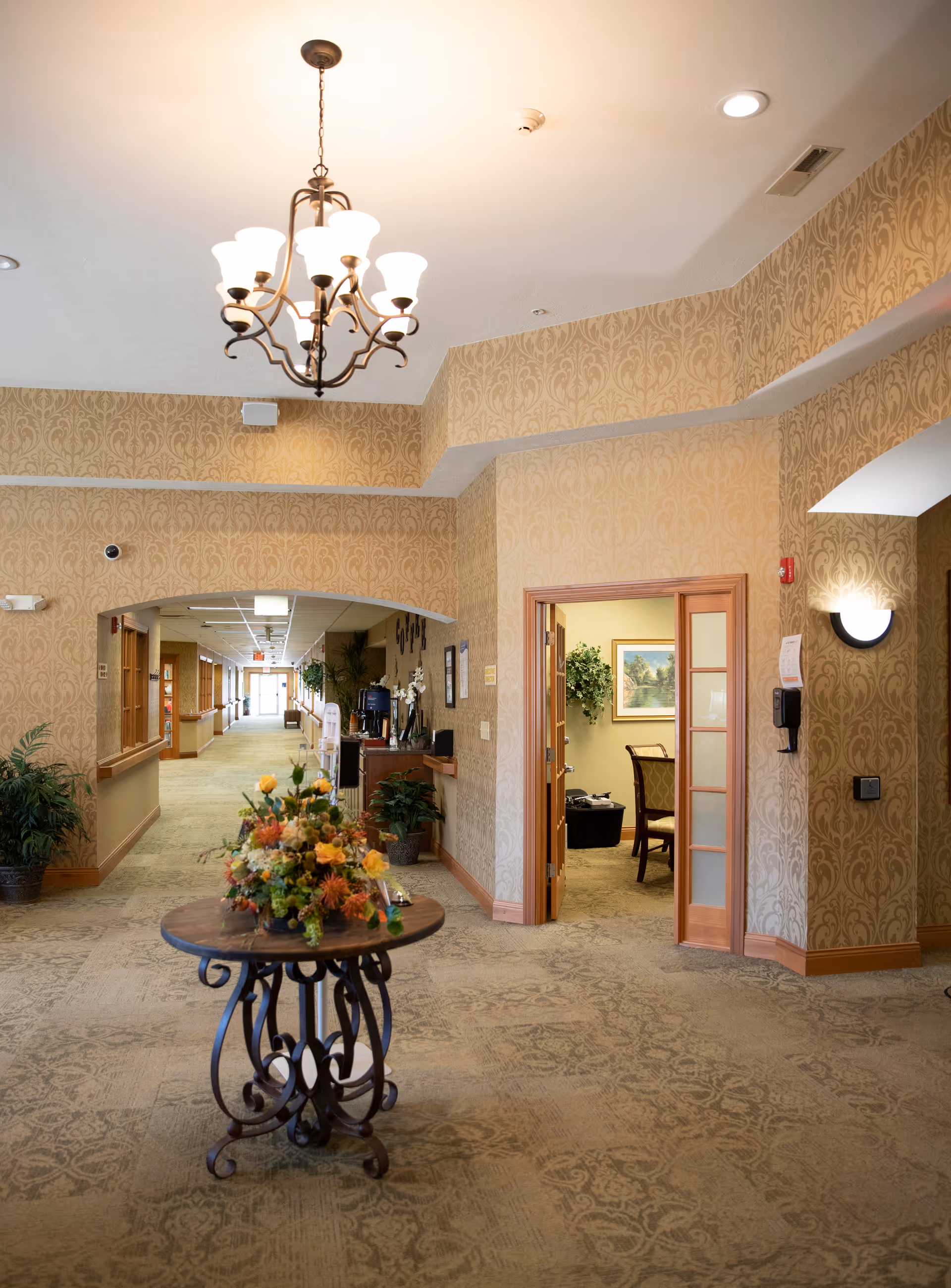 Carpeted reception lobby with a round table holding a floral arrangement, chandelier overhead, a long hallway and an open doorway to a sitting area.