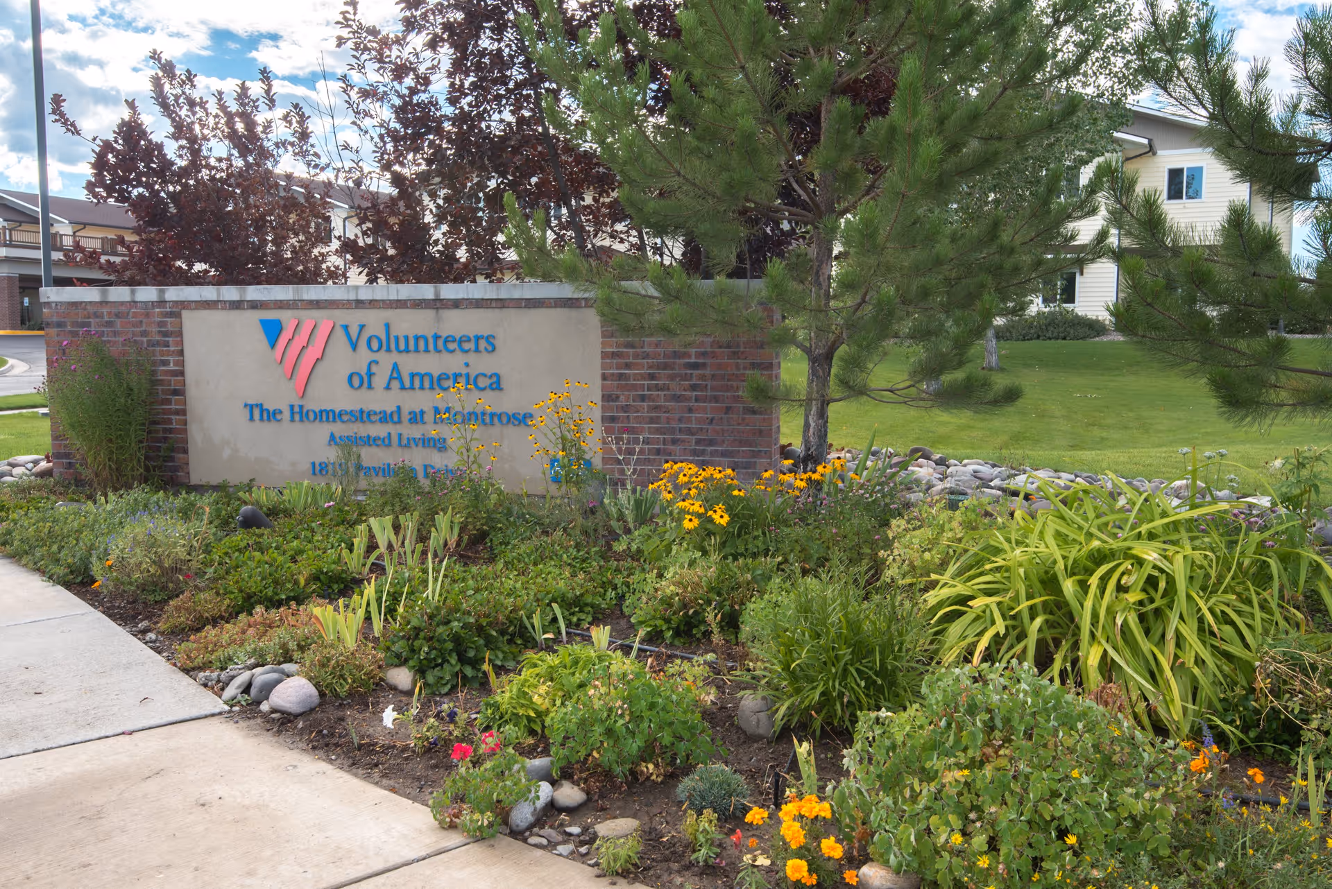 Exterior sign for "Volunteers of America The Homestead at Montrose" surrounded by flowers and landscaping in front of a residential building.