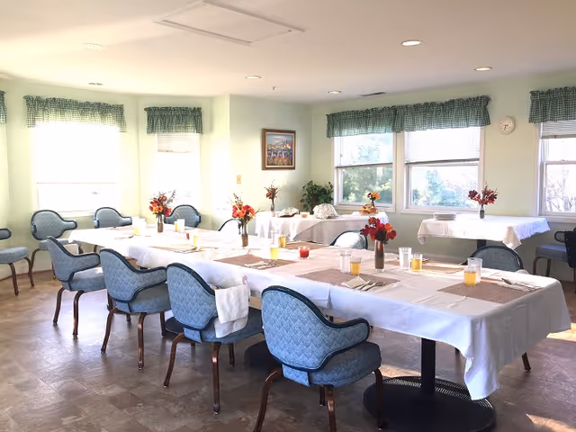 A bright dining room with several tables covered in white tablecloths and set with placemats, glasses, and silverware. Blue upholstered chairs surround the tables. Small vases with red and orange flowers decorate the tables. The room has large windows with green checkered valances, allowing natural light to fill the space. A framed picture and a clock are visible on the walls.