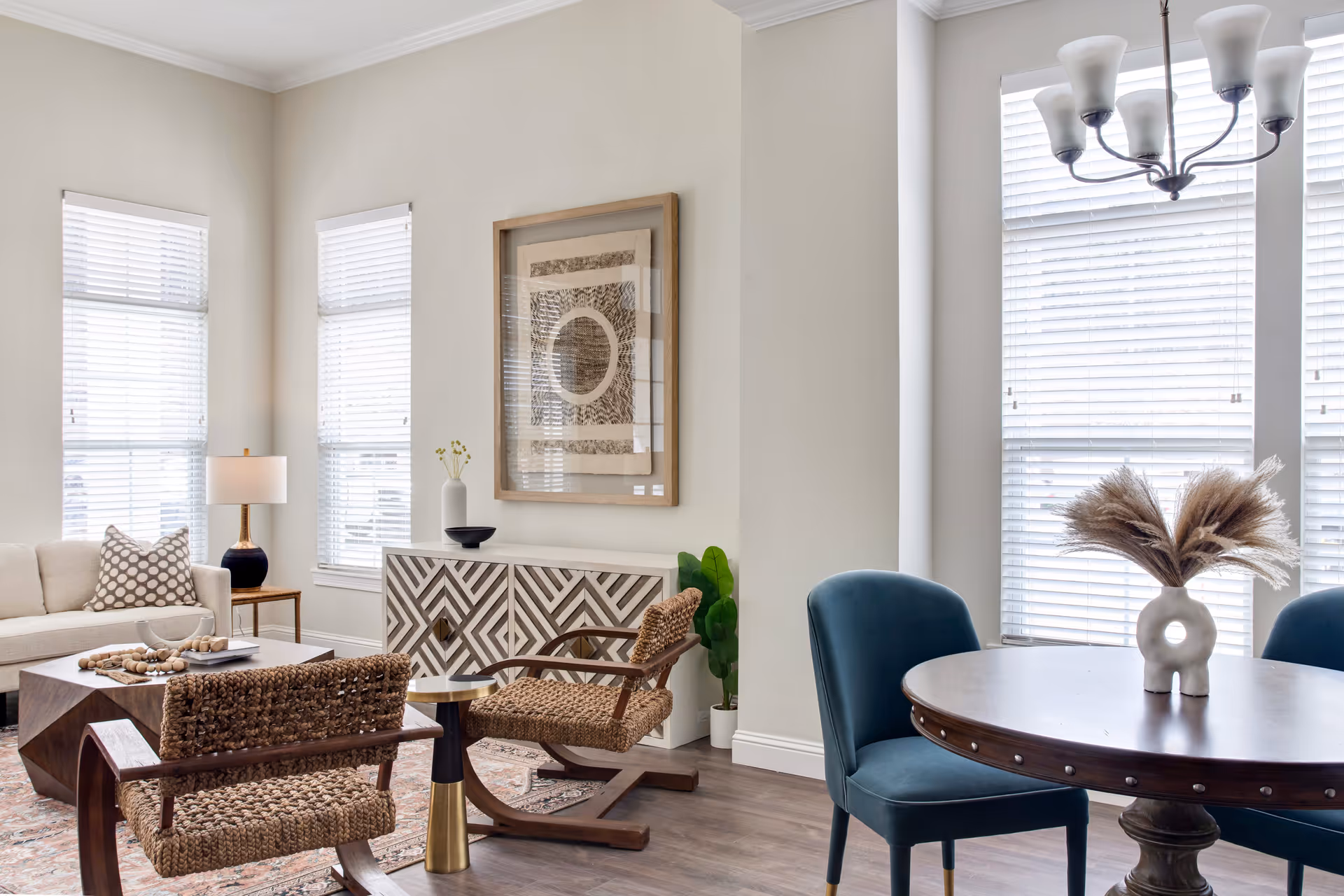 A bright and airy living room with large windows covered by white blinds. The room features a beige sofa with a patterned pillow, two woven armchairs, a wooden coffee table, a patterned sideboard with decorative items, a round wooden dining table with blue upholstered chairs, and a modern chandelier. The walls are light-colored and there is a framed abstract artwork above the sideboard.