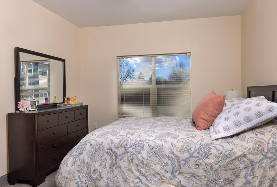 A bedroom with a large window covered by horizontal blinds showing a view of trees and blue sky outside. The room has beige walls and a dark wooden dresser with a large mirror on top. On the dresser are decorative items including a small clock and a coral ornament. A bed with a patterned bedspread in shades of blue and beige is positioned in front of the window, with two pillows, one pink and one white with a floral pattern. A bedside lamp is partially visible behind the pillows.