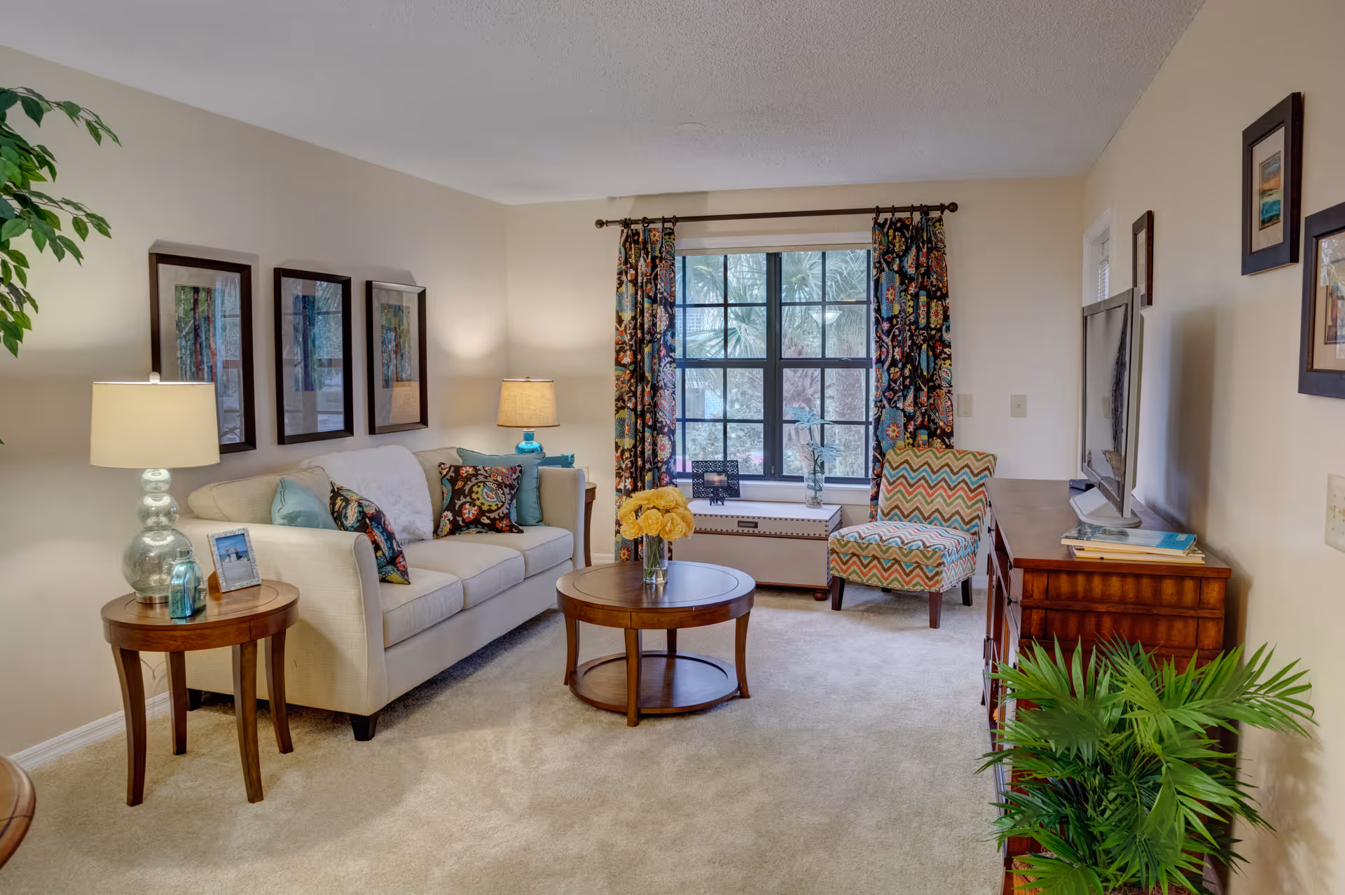 A cozy living room with a beige sofa adorned with colorful patterned pillows, a round wooden coffee table with a vase of yellow flowers, a patterned armchair, and a wooden TV stand with a flat-screen television. The room has a large window with floral curtains, framed artwork on the walls, two table lamps, and a green potted plant in the corner.