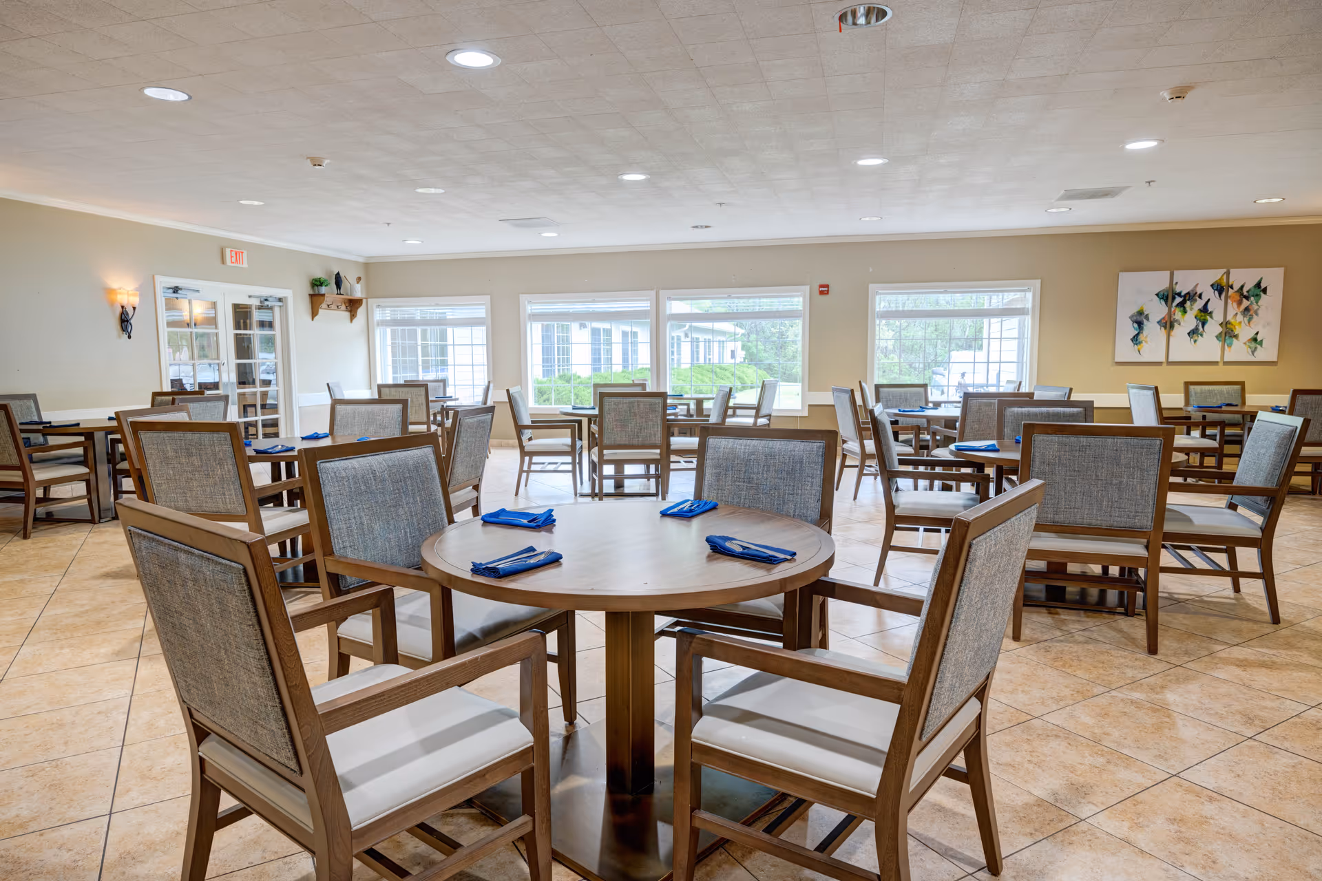 A bright and spacious dining room with multiple round and rectangular wooden tables, each surrounded by cushioned chairs. Blue napkins are neatly placed on the tables. Large windows allow natural light to fill the room, and there is abstract artwork on one wall.