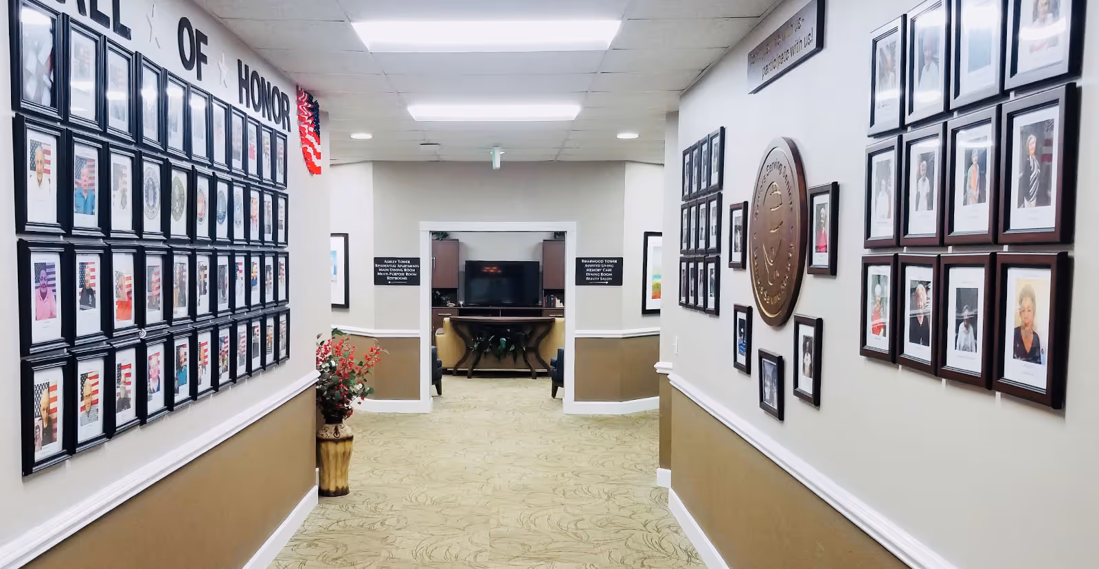 A hallway in a senior living facility with framed photographs on both walls. The left wall features a 'Wall of Honor' with multiple portraits of individuals, each with an American flag background. The right wall has additional framed portraits and a large circular plaque. At the end of the hallway, there is an open doorway leading to a room with a television and chairs. The walls are painted beige and white, and the floor is carpeted with a light pattern. There is a decorative vase with flowers on the left side of the hallway.