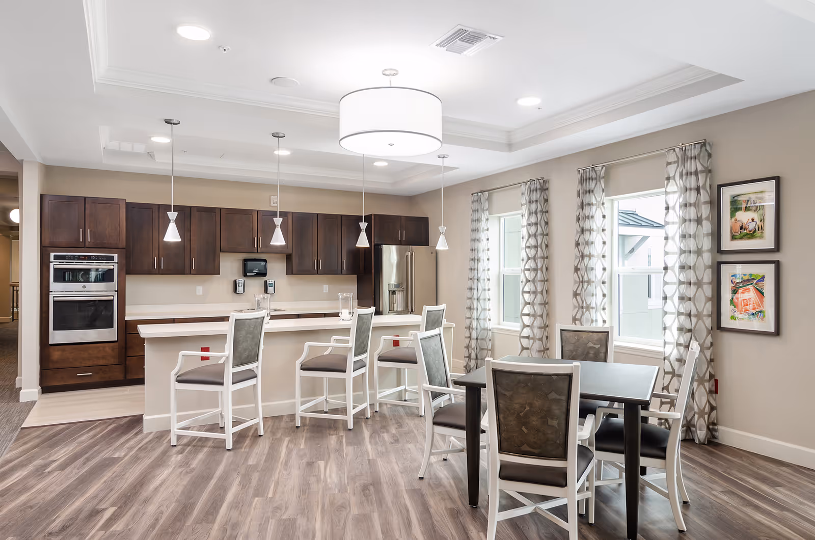 A modern kitchen and dining area in a senior living facility with dark wood cabinets, stainless steel appliances, a kitchen island with four white chairs, a dining table with four chairs, patterned curtains on windows, and framed artwork on the wall.
