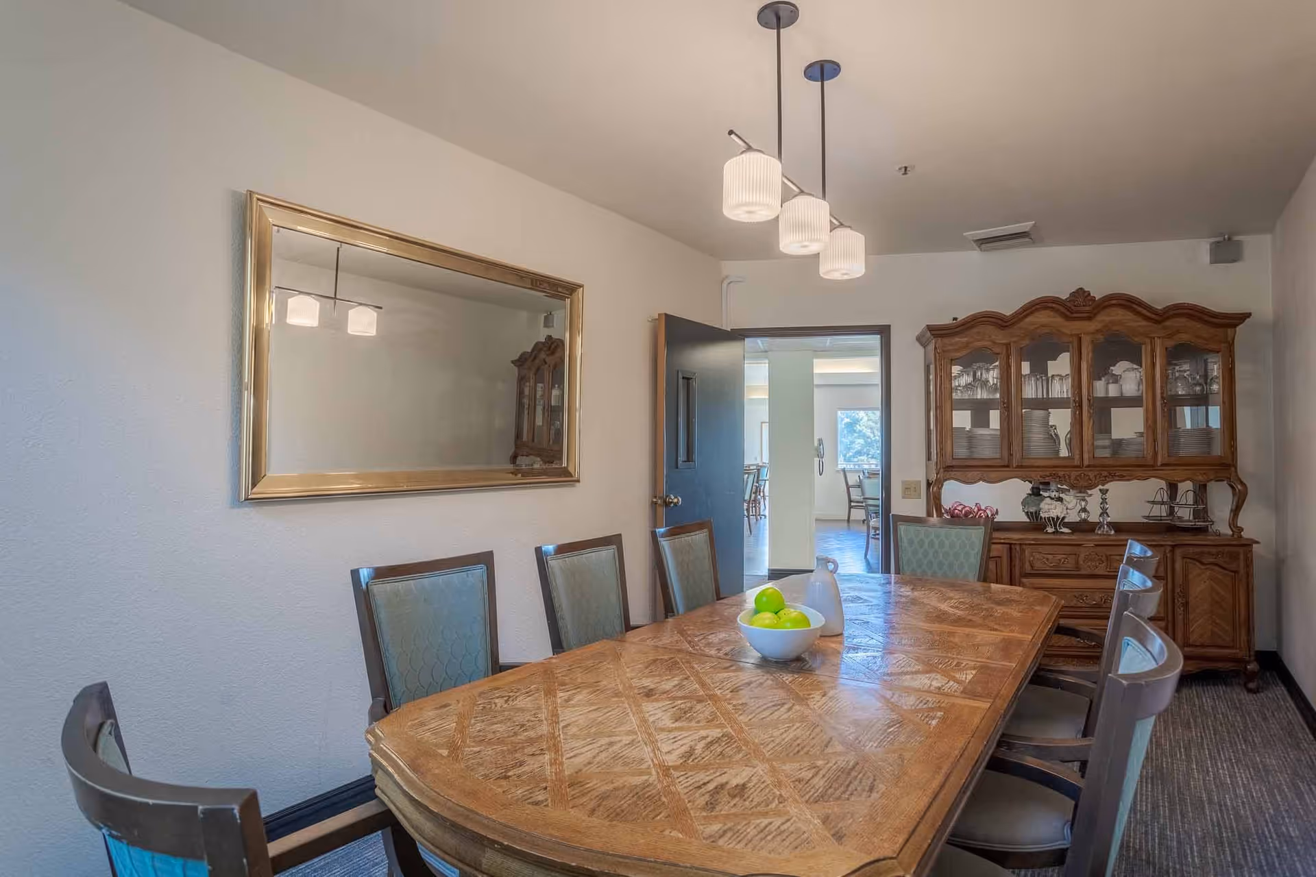 A dining room with a large wooden table surrounded by eight chairs. On the table, there is a bowl with green apples and a white pitcher. A large mirror with a gold frame hangs on the left wall. At the far end of the room, there is a wooden china cabinet with glass doors displaying dishes and glassware. The room is lit by three pendant lights hanging from the ceiling. An open door leads to another room with additional seating and windows.