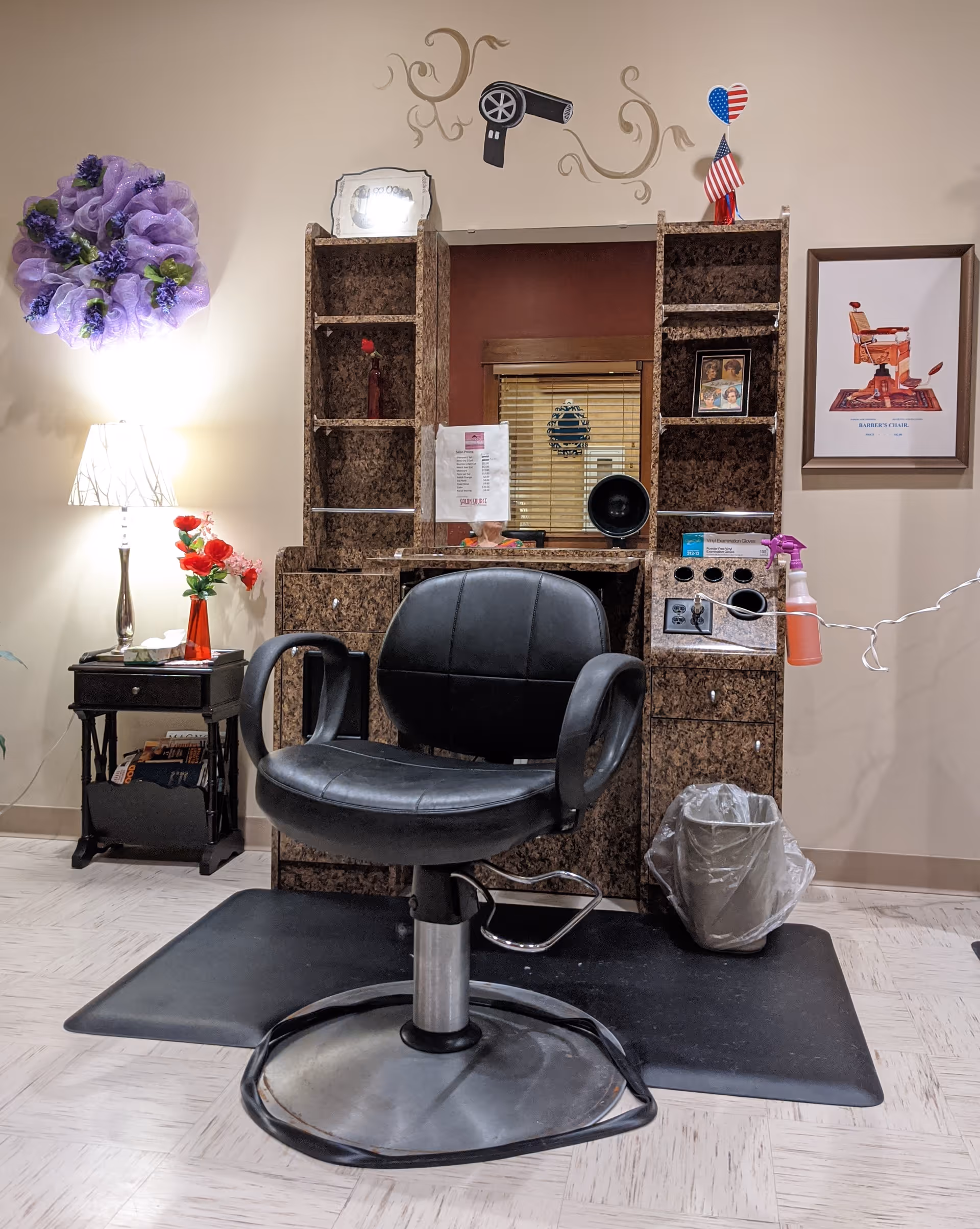 Interior view of a hair salon station with a black salon chair in front of a brown marble-patterned counter and mirror. The wall behind features a decorative hair dryer decal, a framed picture of a barber's chair, and a small American flag decoration. To the left, there is a small table with a lamp and a vase of red flowers, and a purple decorative wreath on the wall.