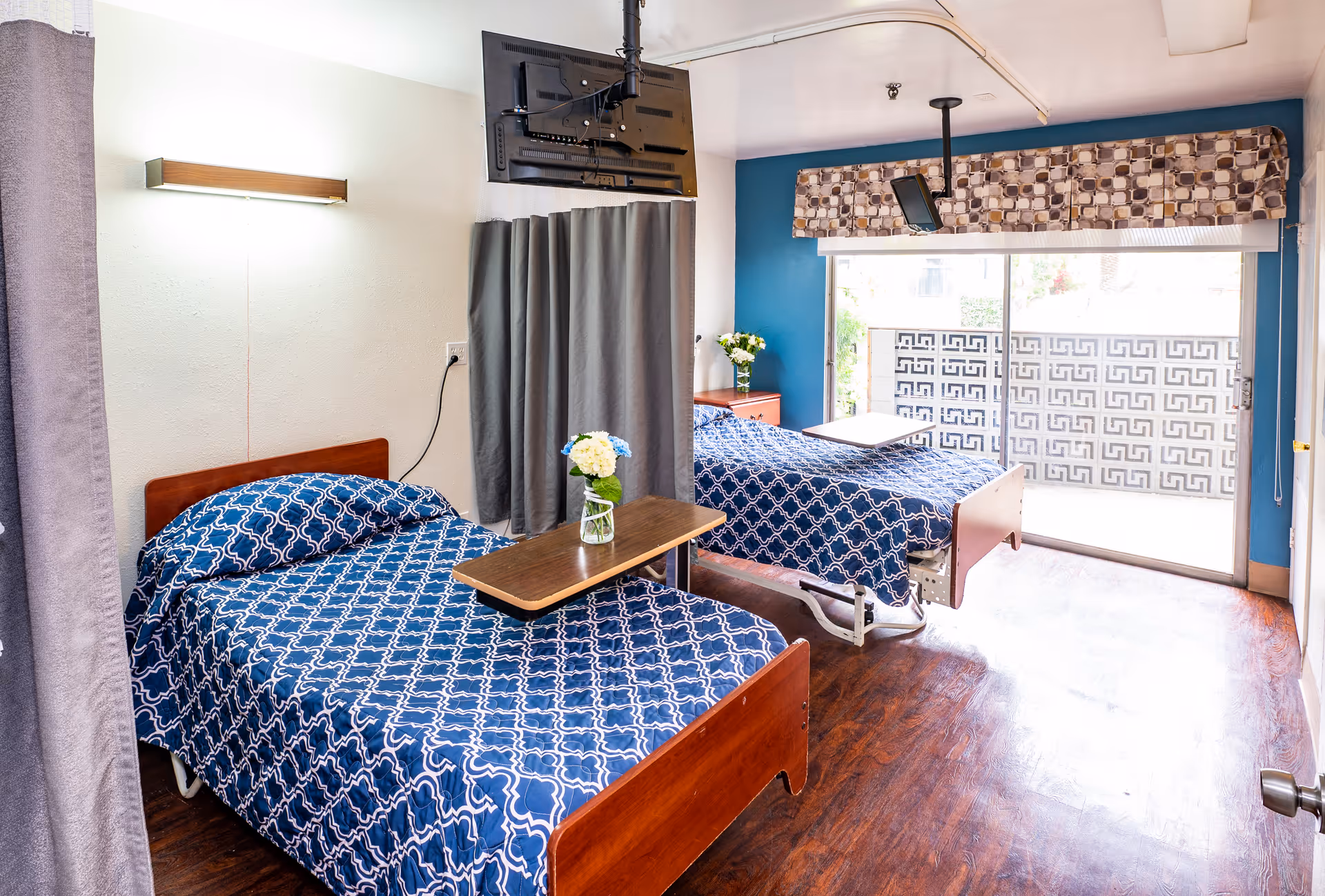 A bright room with two hospital-style beds covered in blue patterned bedspreads. Each bed has a wooden overbed table with a small vase of flowers. A flat-screen TV is mounted on the ceiling between the beds. The room has wooden flooring, a blue accent wall, and a large sliding glass door with patterned curtains leading to an outdoor area with a decorative concrete block wall.