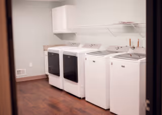 Laundry room with two white washing machines and two white dryers lined up against a light-colored wall with a white cabinet and a wire shelf above them. The floor is dark wood and the room has a clean, organized appearance.