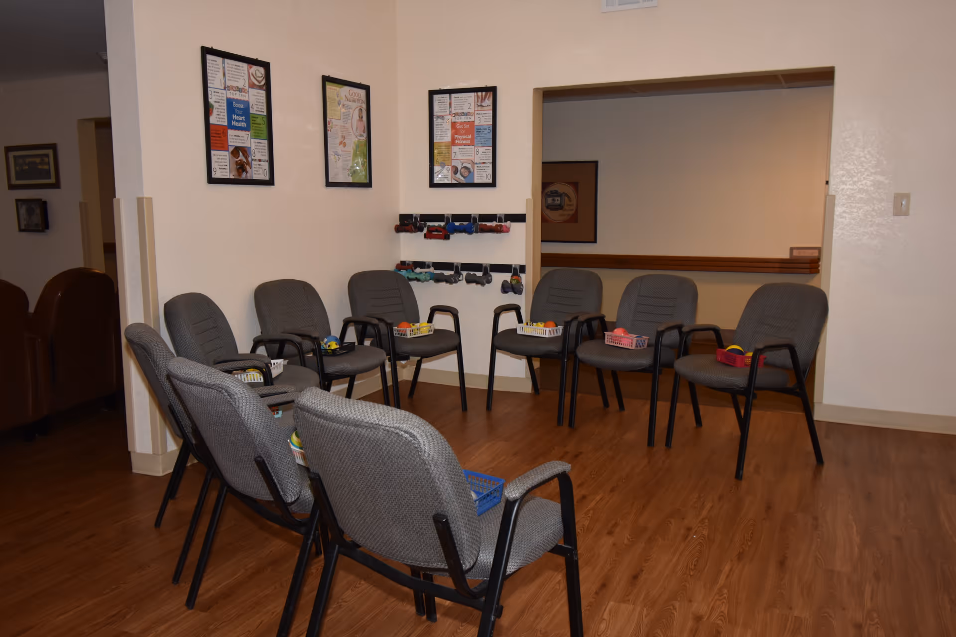 A room with a circle of gray chairs arranged on a wooden floor, each chair holding a small basket with various items. The walls are light-colored and decorated with framed posters and a rack holding small dumbbells.