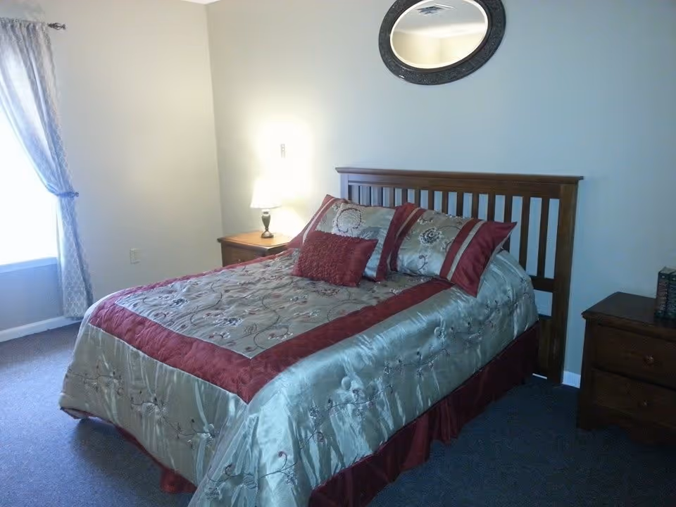 A bedroom with a wooden bed frame and a bed made with a green and burgundy floral patterned comforter and matching pillows. There is a wooden nightstand with a lamp on the left side of the bed and another wooden dresser on the right side with some books on it. A round mirror hangs on the wall above the bed, and a window with patterned curtains is on the left side of the room.