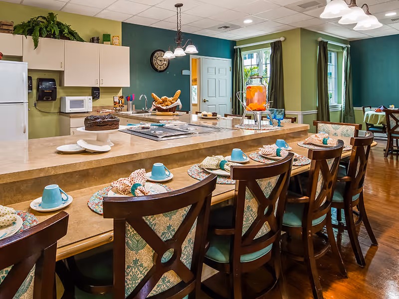 A cozy dining area with a long counter set with plates, cups, and napkins. Behind the counter is a kitchen area with a refrigerator, microwave, sink, and a basket of bread. The room has green walls, wooden chairs with patterned cushions, and windows with green curtains letting in natural light.