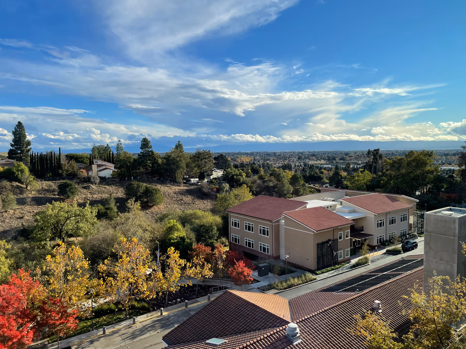 Aerial view of a retirement community building with red-tile roofs, surrounding trees and landscaping, and a distant city skyline under a partly cloudy blue sky.