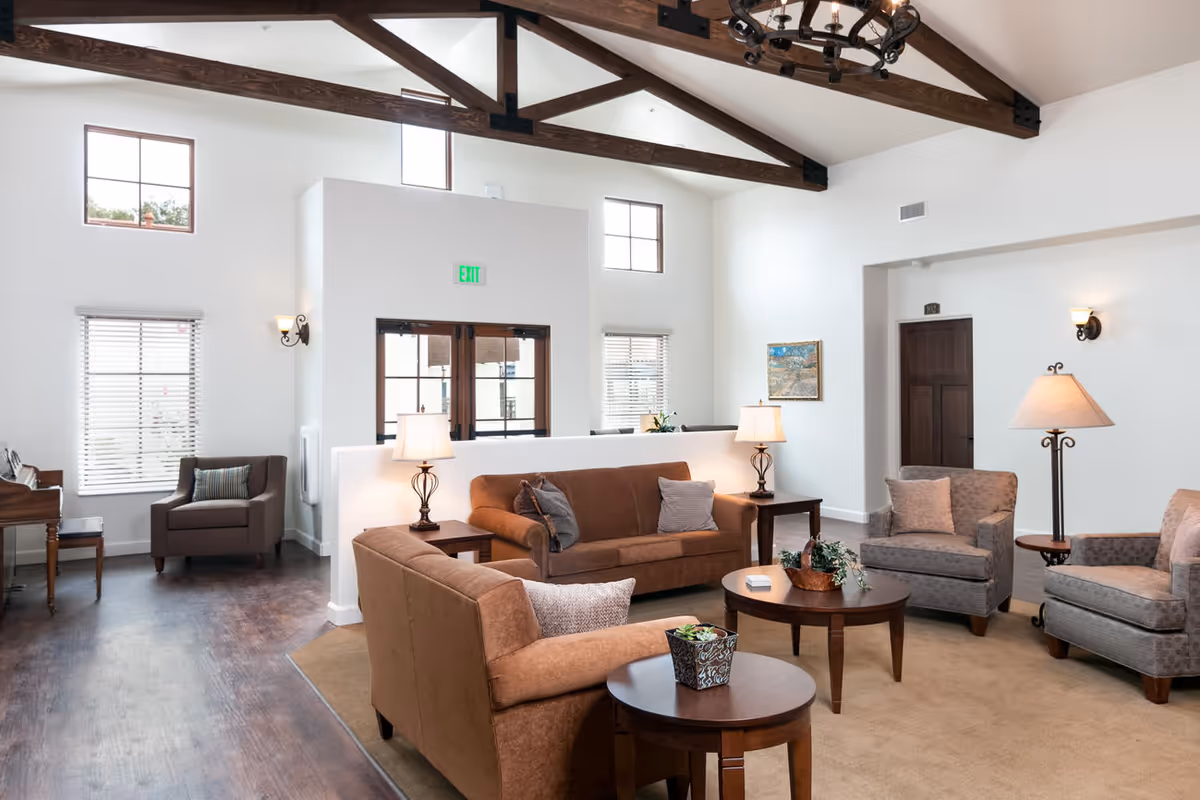 A bright and spacious living room with high vaulted ceilings featuring exposed wooden beams. The room is furnished with brown and gray upholstered sofas and armchairs arranged around wooden coffee and side tables. There are table lamps on the side tables and wall sconces providing warm lighting. Windows with blinds allow natural light to enter, and a piano is visible in the corner near one window. The walls are white, and there is a painting hanging on one wall. The floor is a combination of dark wood and a beige carpet.