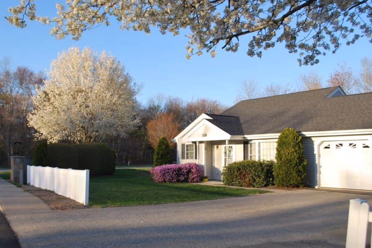 A single-story residential building with a white exterior and a dark roof, surrounded by green bushes and flowering trees. There is a white picket fence along the driveway and a clear blue sky above.