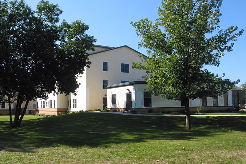 Exterior view of a multi-story senior living facility building with light-colored siding, surrounded by green grass and trees under a clear blue sky.
