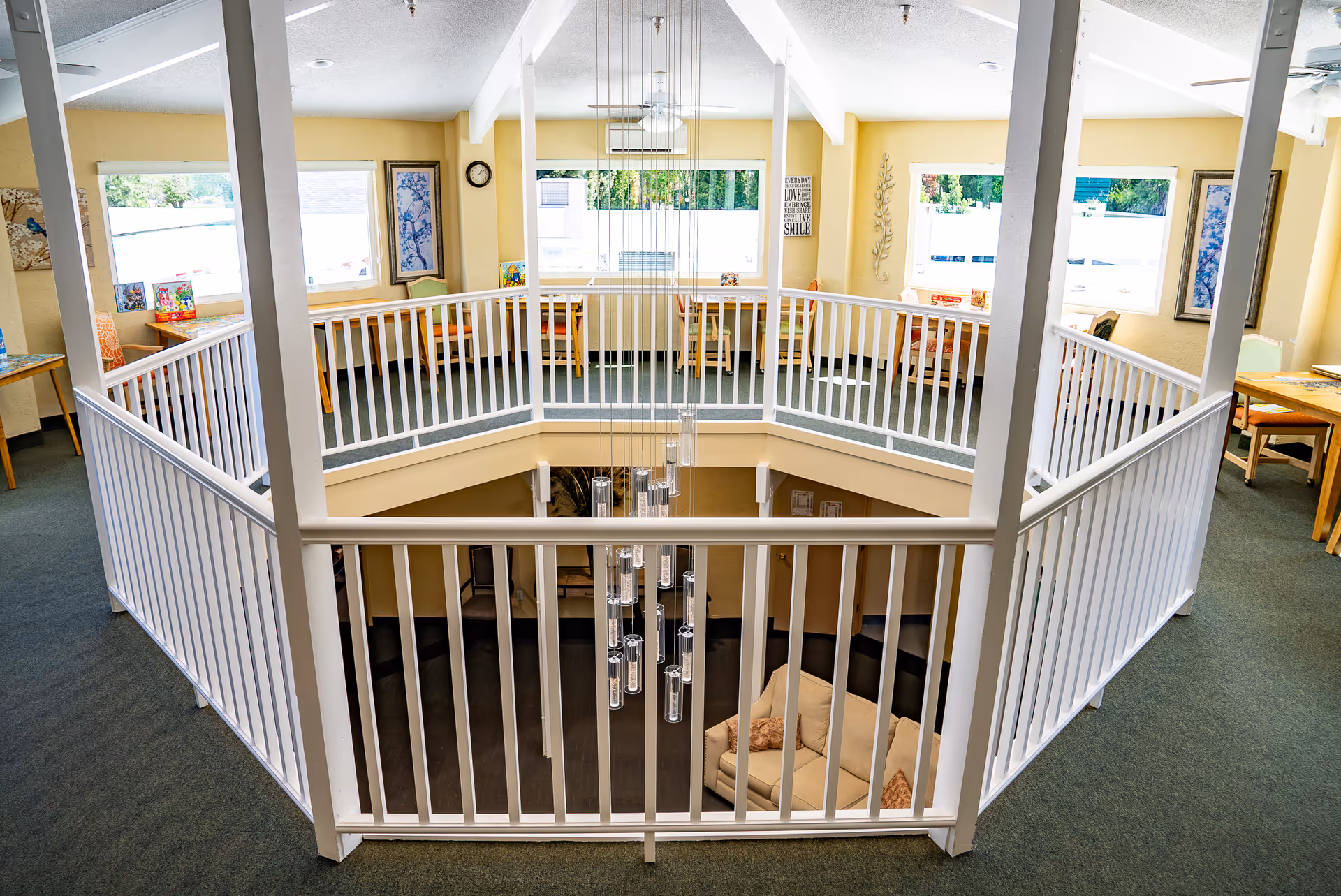 Interior view of a senior living facility showing a second-floor balcony with white railings overlooking a lower level with a beige sofa. The upper floor has several tables and chairs near windows with natural light coming in. The walls are painted yellow and decorated with framed artwork and wall hangings. A modern chandelier hangs in the center.