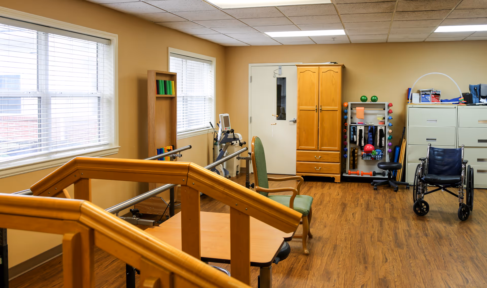A rehabilitation room with wooden handrails for walking exercises, a green cushioned chair, a wooden cabinet, exercise equipment including weights and therapy balls, a wheelchair, and filing cabinets. The room has beige walls, two windows with blinds, and a wooden floor.