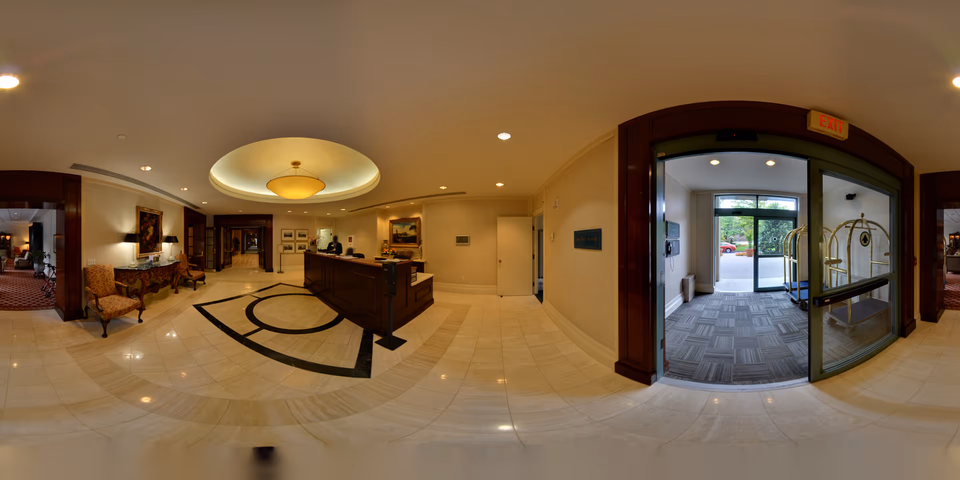 A spacious and well-lit reception area in a senior living facility with a wooden front desk, a receptionist, elegant chairs, artwork on the walls, and a glass entrance door leading outside. The floor is tiled with a decorative pattern, and the ceiling features a large circular light fixture.