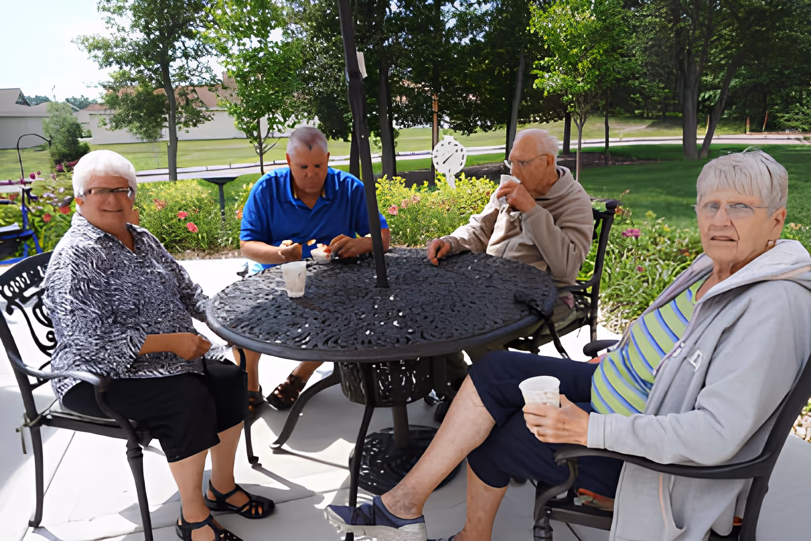 Four elderly people sitting around a black metal outdoor table with an umbrella, enjoying drinks and snacks in a garden area with green grass, trees, and flowers in the background.