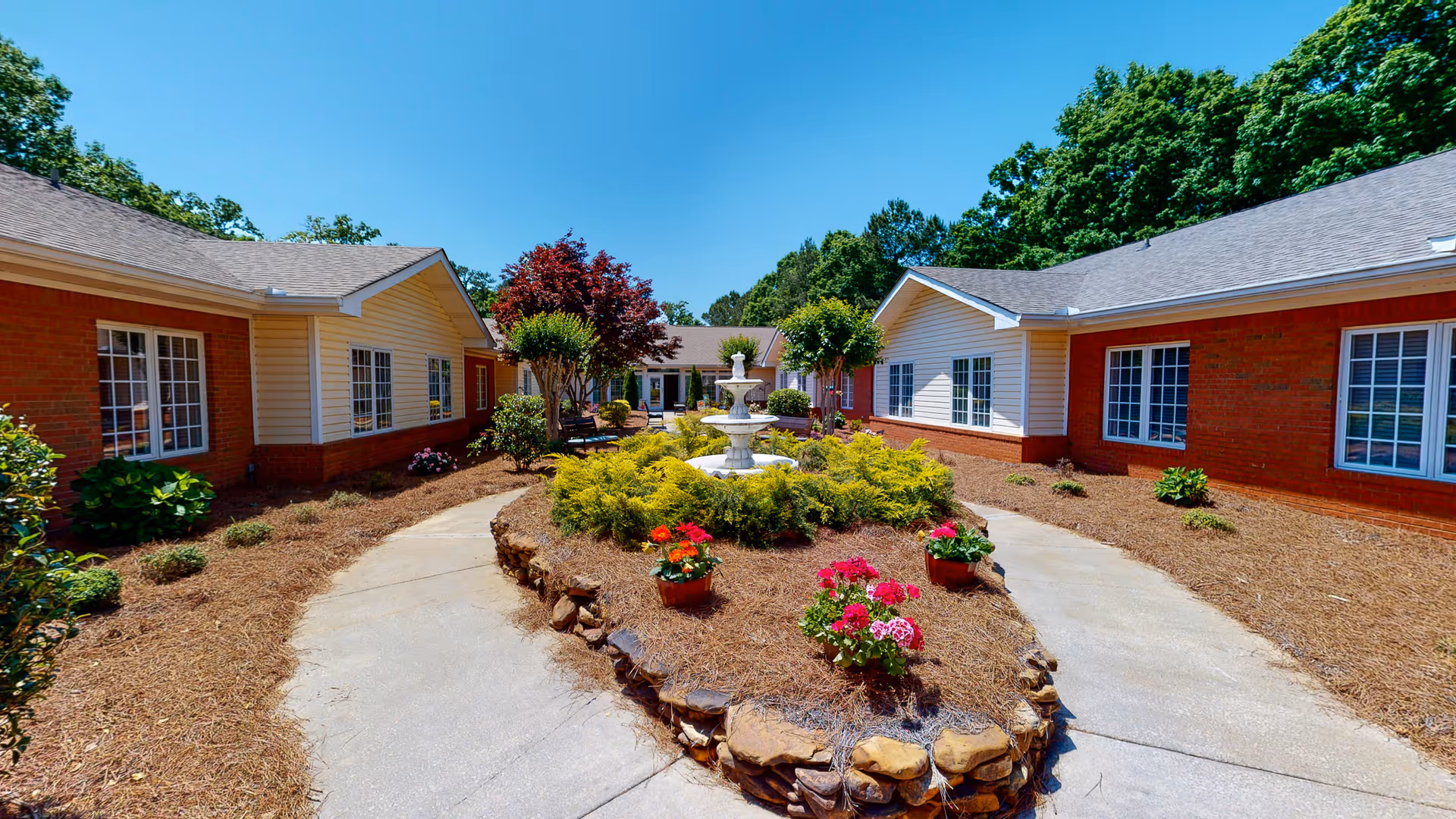 Sunny landscaped courtyard between single-story red-brick buildings with a central fountain and flowerbeds.