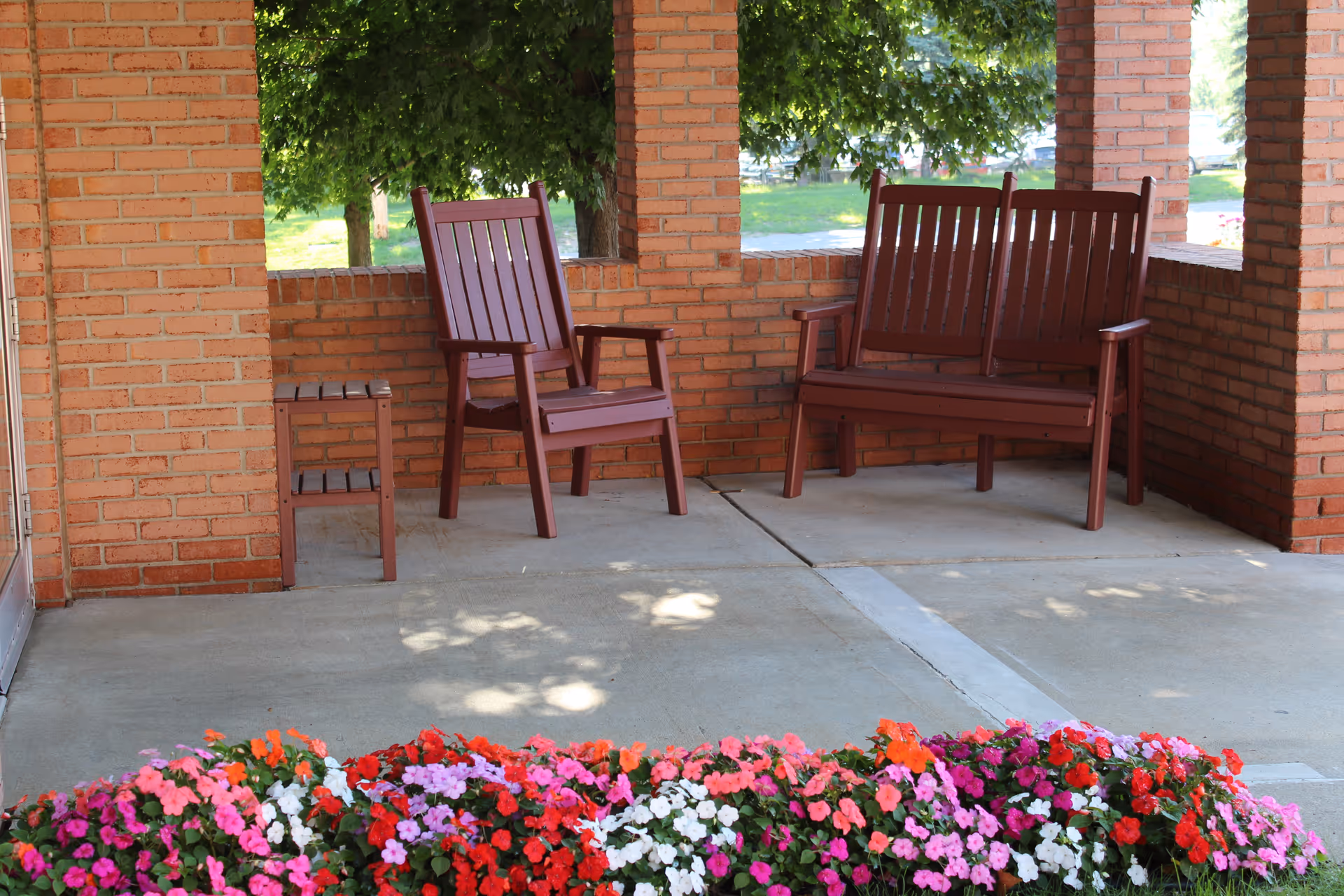 Covered brick porch featuring red wooden chairs and a bench overlooking a colorful flower bed.
