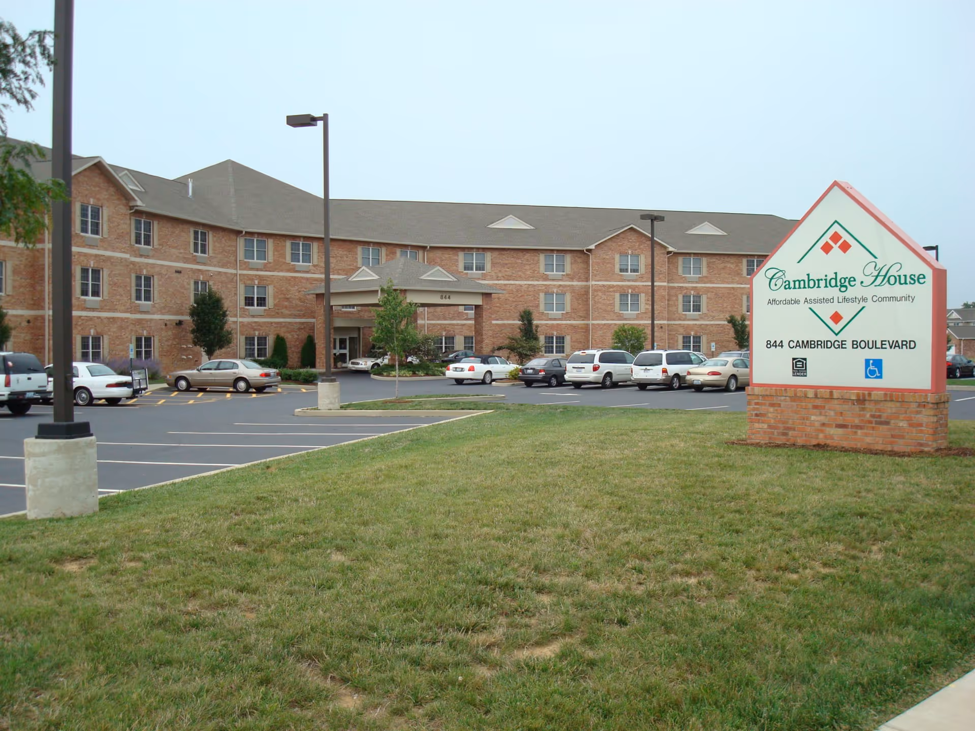 Exterior view of Cambridge House of Ofallon, an affordable assisted lifestyle community. The image shows a large three-story brick building with multiple windows, a covered entrance, a parking lot with several cars, and a sign displaying the facility's name and address at 844 Cambridge Boulevard.
