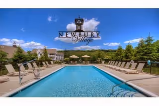 Outdoor swimming pool with lounge chairs arranged on both sides under a clear blue sky with some clouds. Trees and buildings are visible in the background. The image has the text 'Newbury Village' overlaid at the top center.