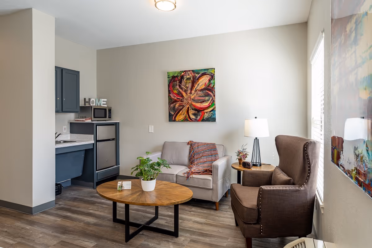 Bright living room with a small kitchenette, a sofa and armchair around a round coffee table, and colorful wall art.