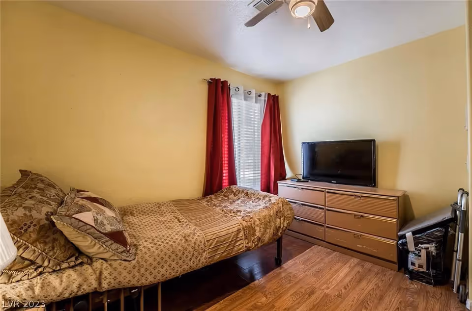 A small bedroom with a single bed covered in patterned brown bedding and pillows. There is a window with white blinds and red curtains. A wooden dresser with a flat-screen TV on top is positioned against the wall. The room has light yellow walls, wood flooring, and a ceiling fan with a light.