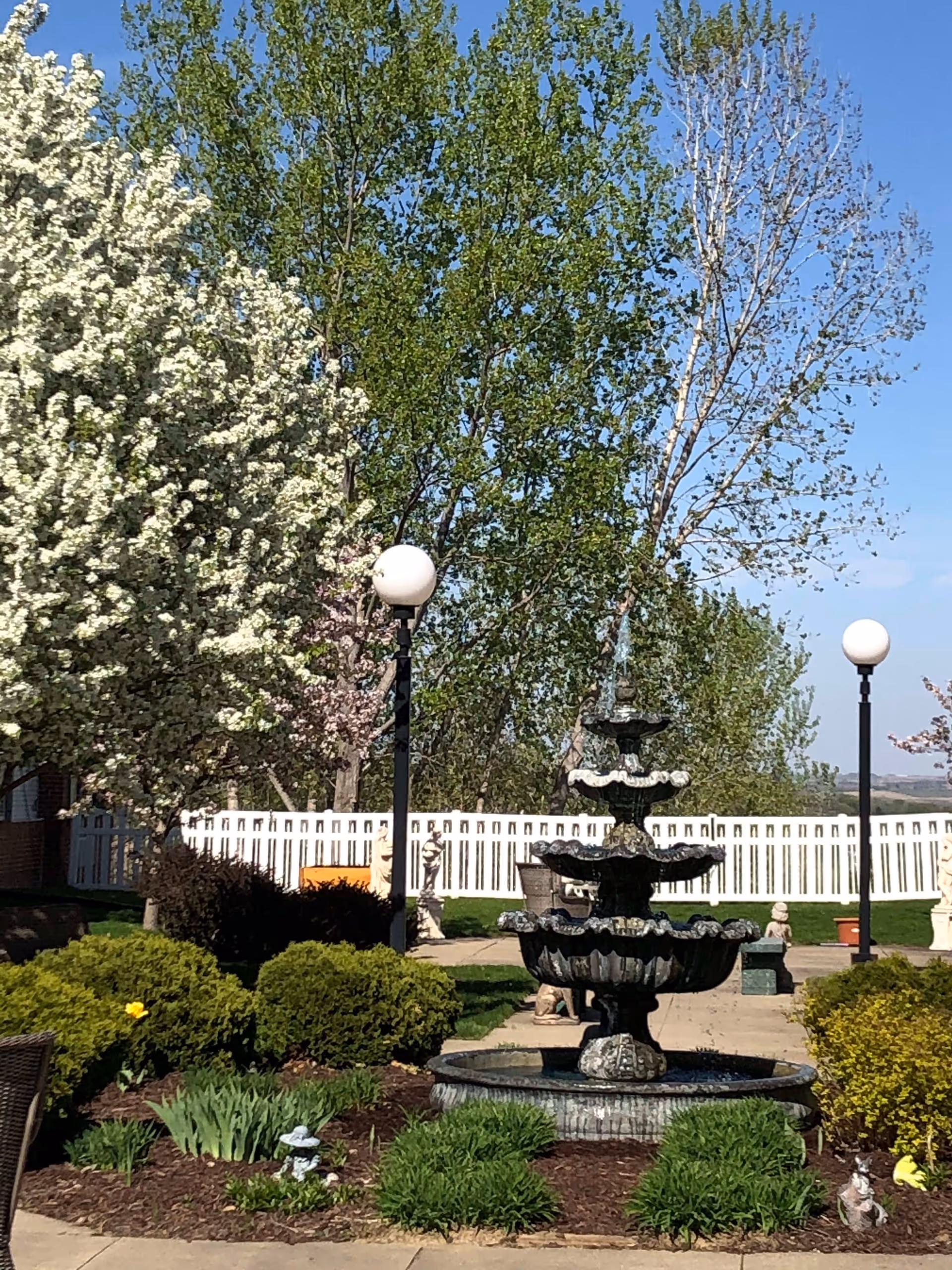 Outdoor garden area with a multi-tiered stone fountain in the center, surrounded by green bushes and blooming trees. There are two black lamp posts with round white lights, a white picket fence in the background, and a clear blue sky.