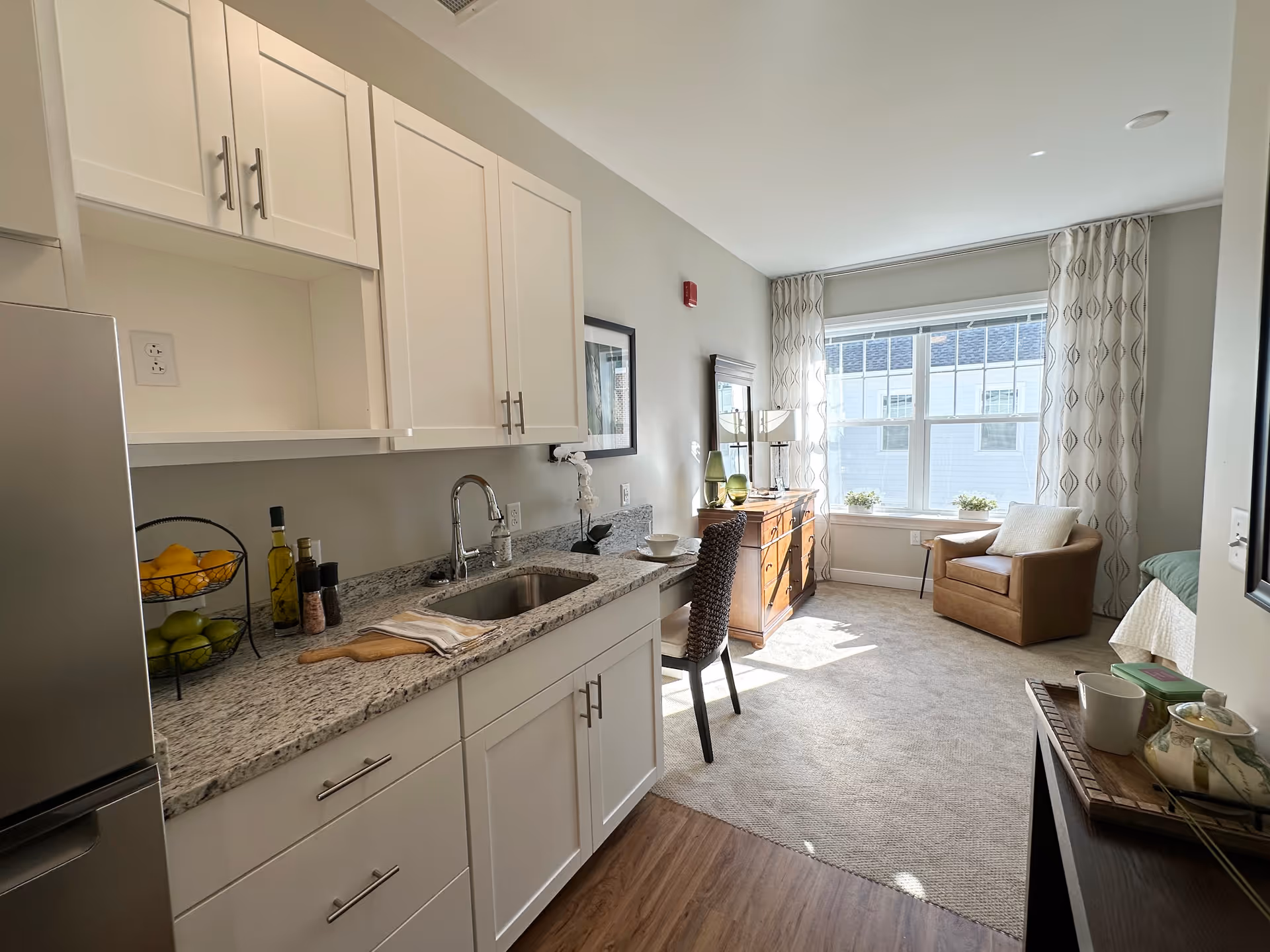 A bright and cozy studio apartment with a small kitchen area featuring white cabinets, a granite countertop, a stainless steel sink, and a refrigerator. The living space includes a wooden dresser, a comfortable armchair near a large window with patterned curtains, and a small desk with a chair. The room is carpeted with sunlight streaming in through the window.